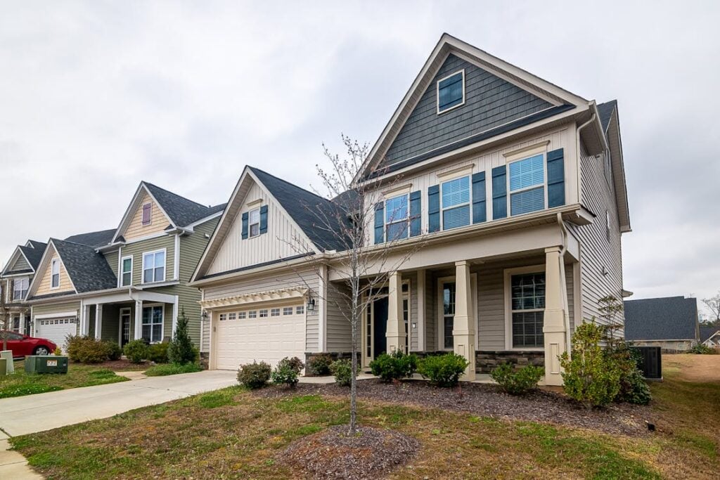 A row of suburban two-story houses with front porches and garages, set against a cloudy sky. The homes, evocative of a working-class neighborhood, are situated along a concrete driveway with minimal landscaping.