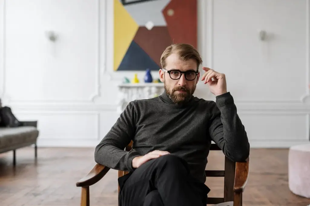 A man with glasses sits in a wooden chair in a modern room with abstract art on the wall.