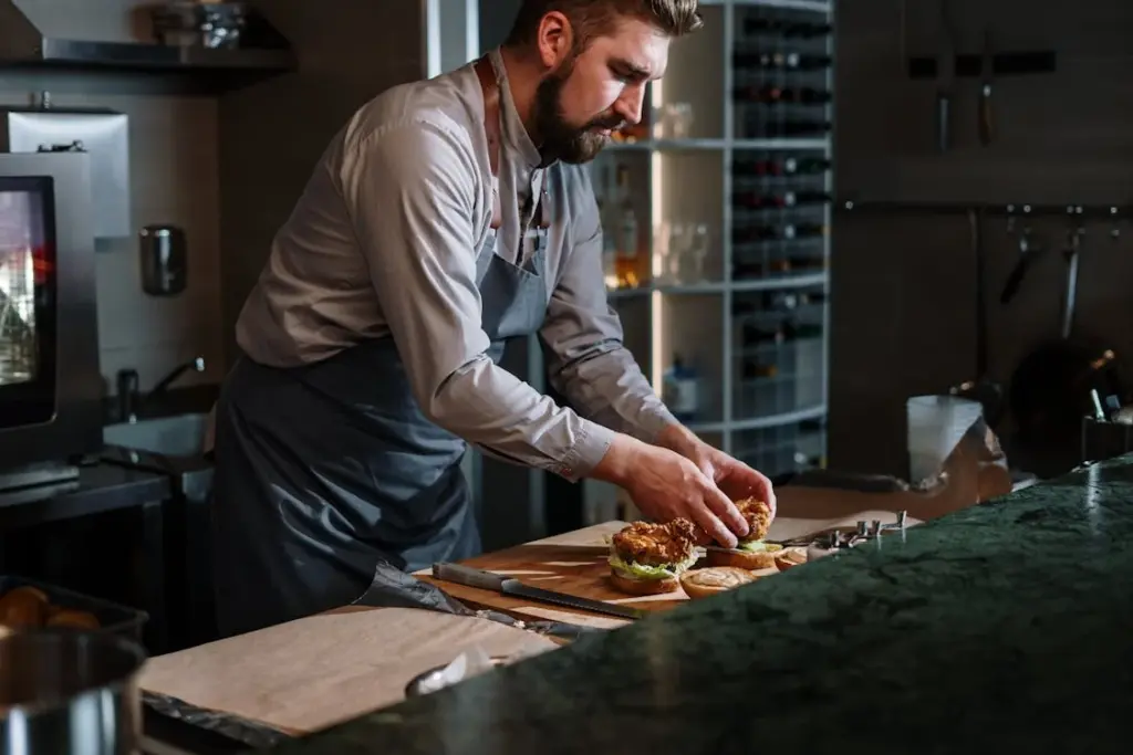 A chef assembles a sandwich at a kitchen counter, surrounded by cooking utensils, with a focused expression.