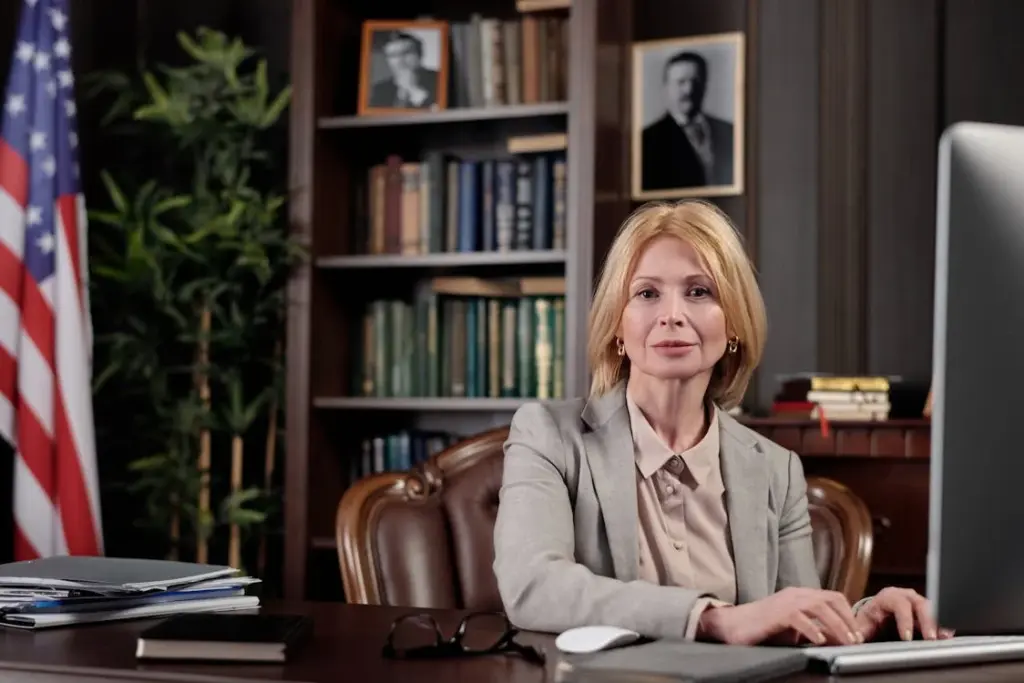 A woman in a beige blazer sits at a desk with a computer in an office. An American flag, bookshelf, and framed photos are in the background.