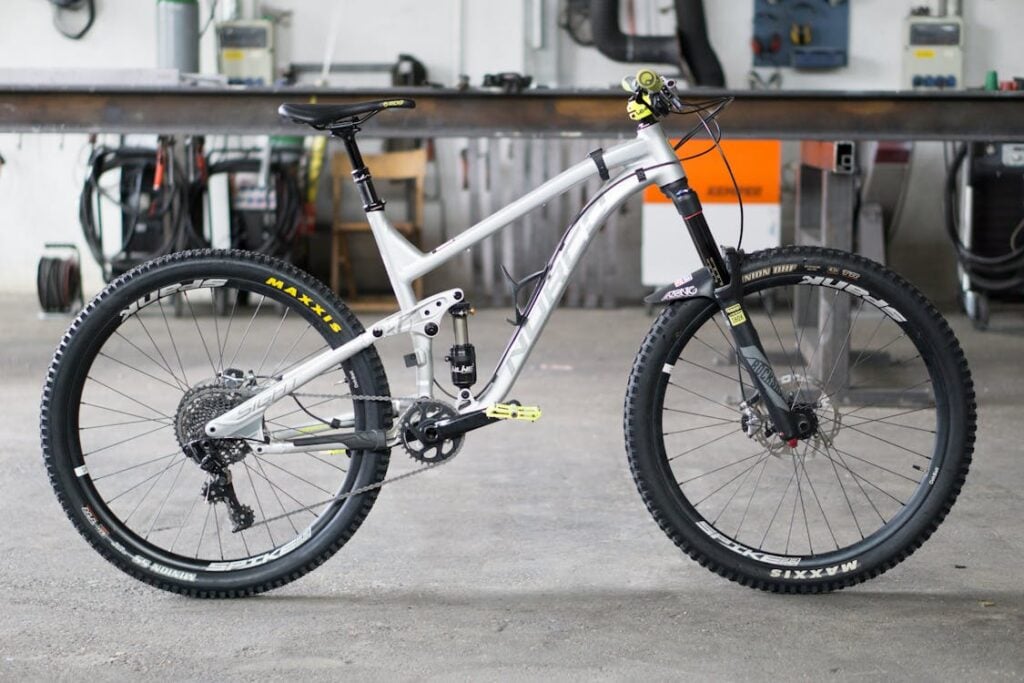 A silver mountain bike with black tires and yellow detailing, resting on a concrete floor in a workshop with various tools and equipment in the background.