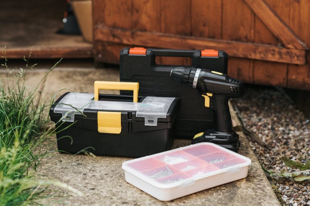 A set of tools, including a toolbox, a drill, and a case with assorted screws, is placed on a concrete surface in front of a wooden shed.