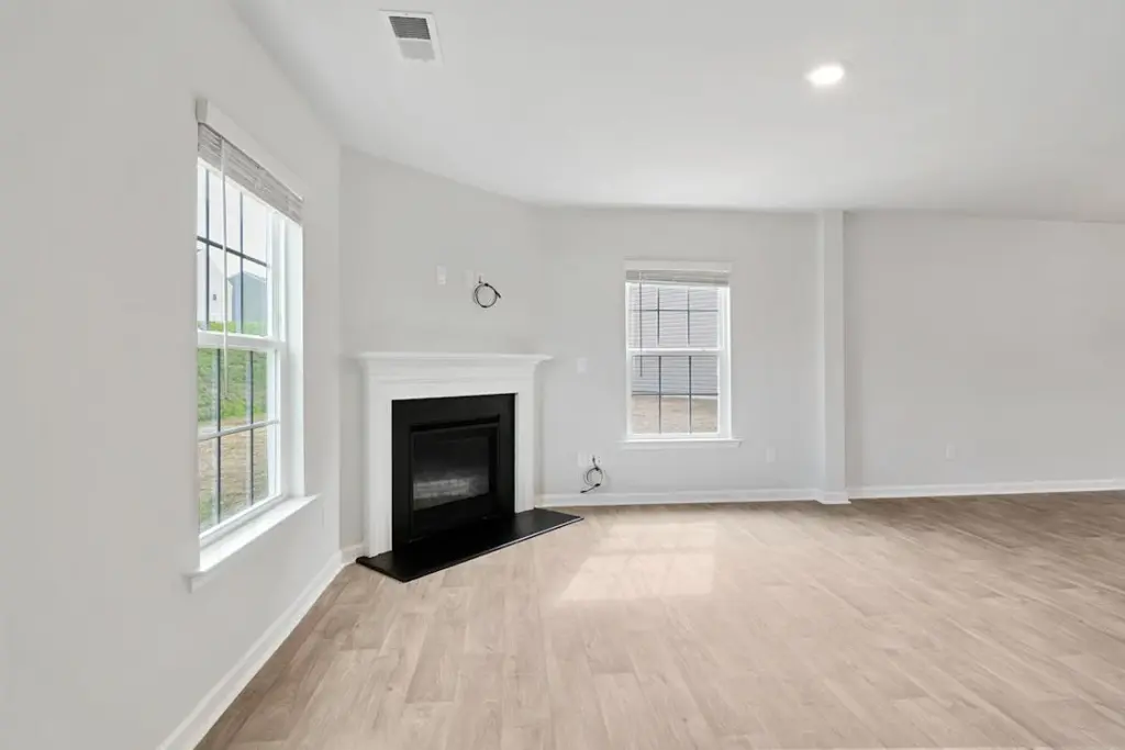 A bright, empty room with light-colored hardwood flooring, two windows with blinds, and a white fireplace. The walls are painted white, and there is recessed lighting in the ceiling.