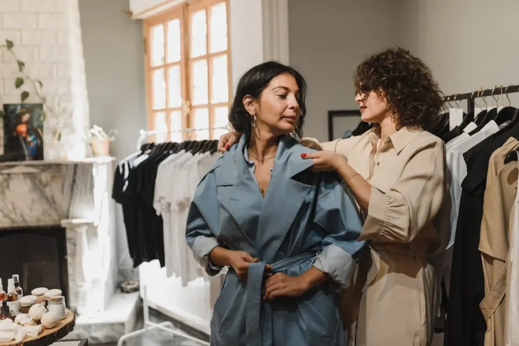 Two women in a clothing store; one, likely from an upper middle class income bracket, is wearing a blue coat while the other adjusts it. Racks of clothes are visible in the background.