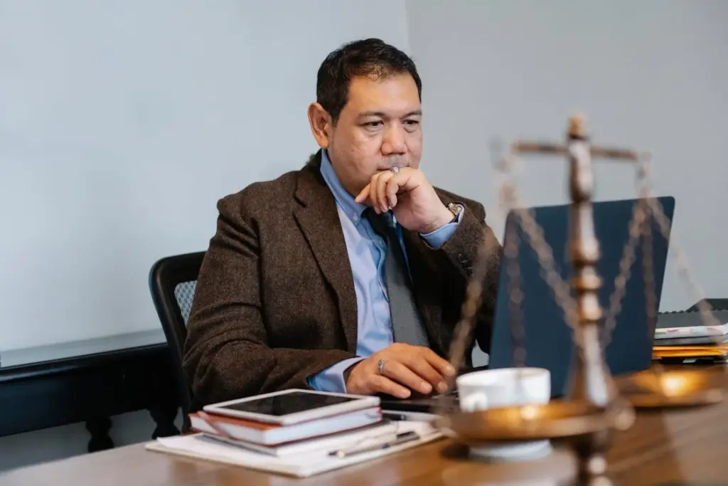 A man in business attire sits at a desk working on a laptop, with legal scales, a tablet, notebooks, and a cup of coffee in the foreground.