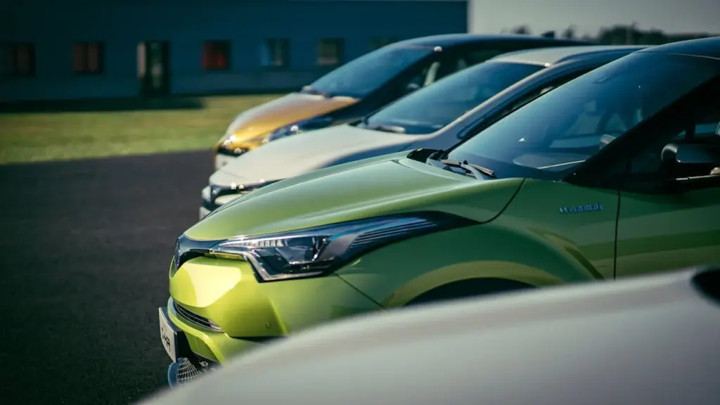 A row of parked cars, primarily in shades of green and white, are aligned on an asphalt surface near a building with windows, suggesting a neighborhood characterized by lower middle class income.