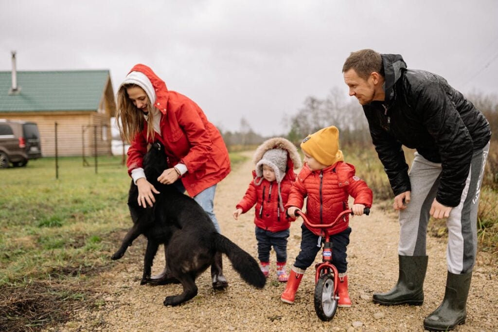 A family of four with two young children and a black dog are outside on a gravel path near a house. One child is on a balance bike, and both adults are wearing jackets and boots.