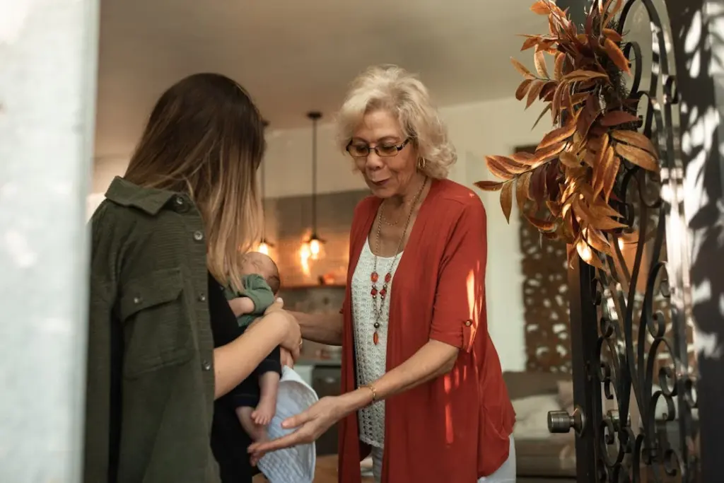 A woman wearing glasses and a red cardigan greets another woman holding a baby at the door.