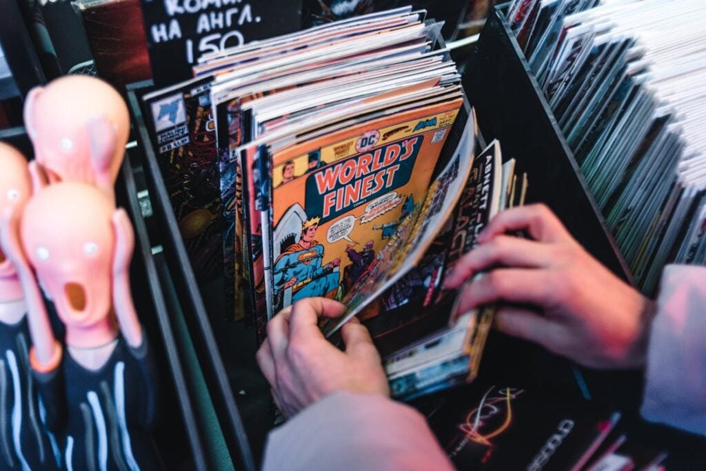 A person flips through a collection of comic books in a store, with figurines and price tags visible in the background.