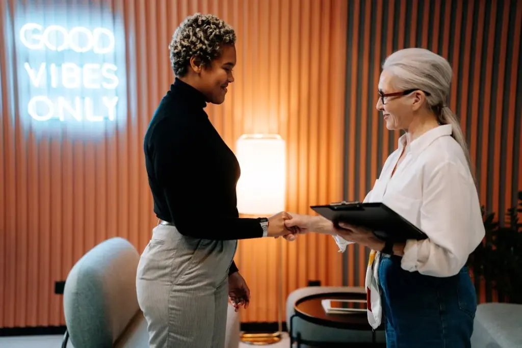 Two women are shaking hands in a modern office setting with a neon sign that reads 