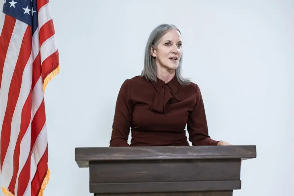 A woman stands at a wooden podium with an American flag to her left, speaking against a plain white background.