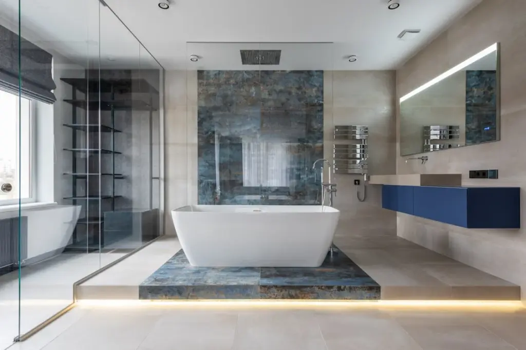 Modern bathroom with a central white bathtub placed on a raised platform, large mirror, floating vanity with blue cabinets, walk-in glass shower, and beige tiled walls and floor.