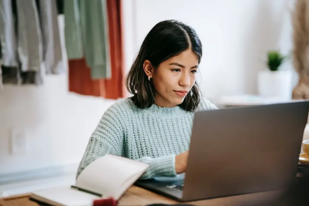 A woman with shoulder-length hair is looking at a laptop while sitting at a table. A notebook is open in front of her. She is wearing a light blue sweater.