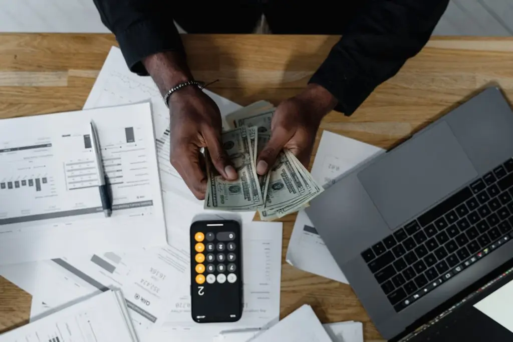 A person counts dollar bills at a desk with financial documents, a calculator, and a laptop.