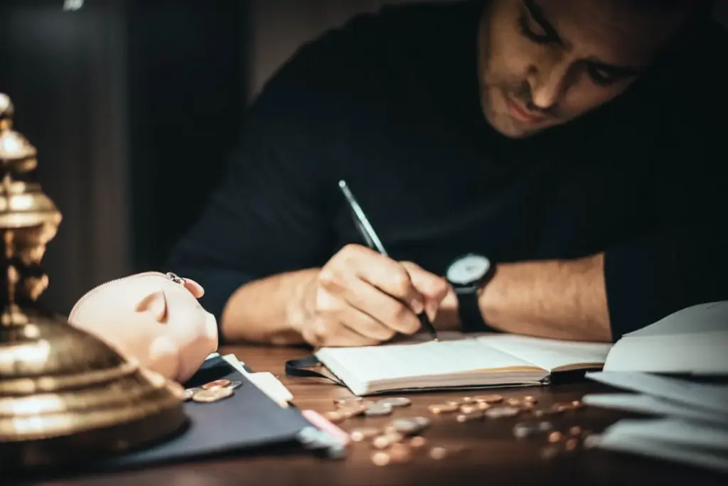 A person writes in a notebook beside coins, a piggy bank, and a lamp on a wooden table.