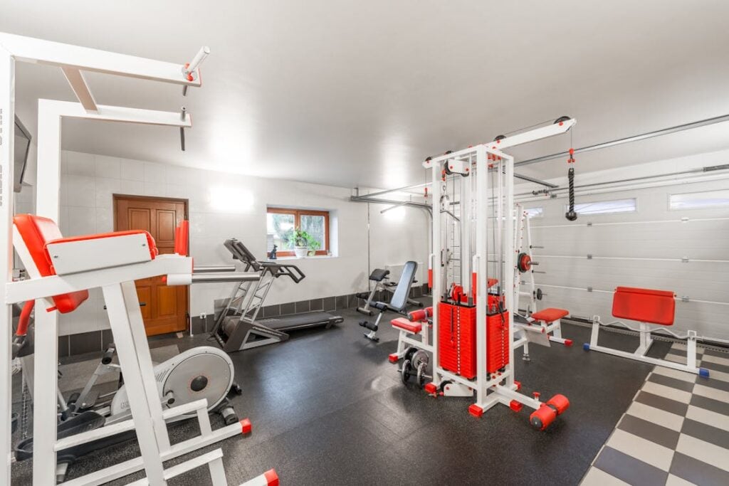 A home gym with various workout equipment, including a treadmill, exercise bike, weight machine, and bench press, arranged in a room with white walls and a checkered floor.