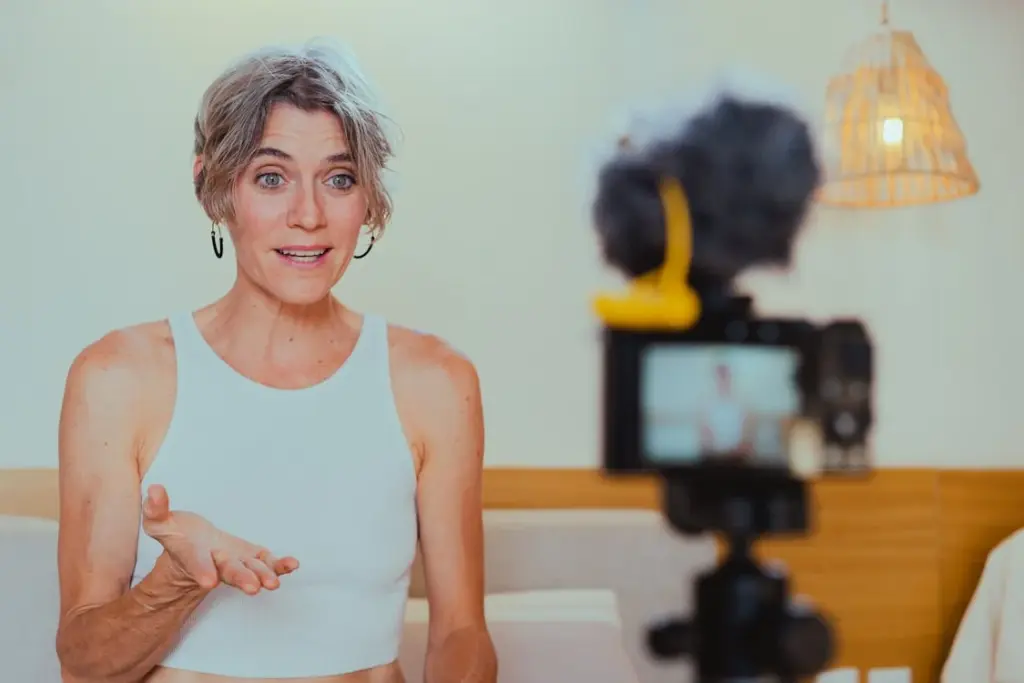 A person with short gray hair wearing a white sleeveless top is speaking in front of a camera setup with a microphone. They are inside a room with a white wall and a hanging rattan lamp.