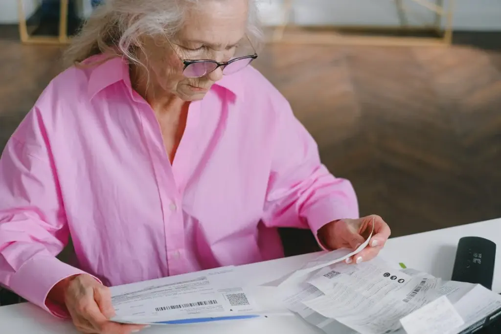 Elderly woman wearing glasses and a pink shirt sits at a table looking at several pieces of paperwork and receipts.