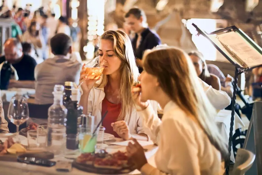 Two women sit at a table in a busy restaurant, one drinking a beverage and the other eating. Various dishes and drinks are on the table. Other patrons are visible in the background.