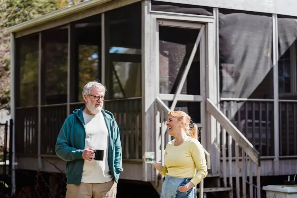 Two elderly individuals, one wearing a green hoodie and the other in a yellow sweater, are standing outside a screened porch, holding mugs and engaging in conversation.