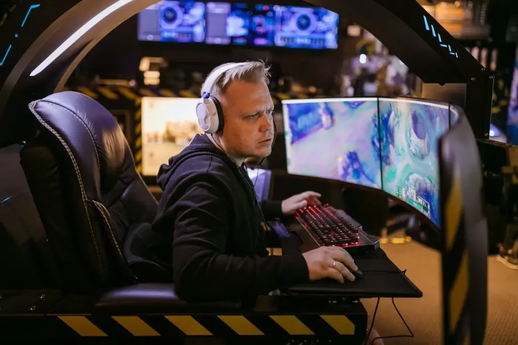 A person wearing headphones is sitting in a gaming chair, using a computer with three curved monitors displaying a video game. The background includes additional screens and gaming equipment.