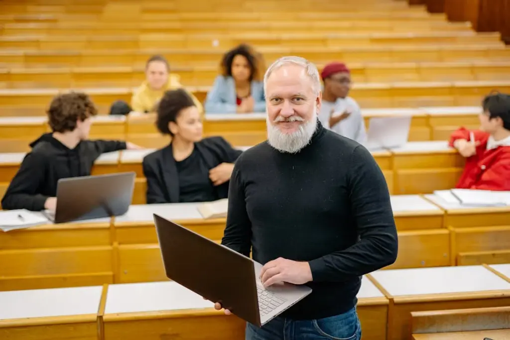 A bearded man stands with a laptop in a lecture hall, with several students seated and engaged in discussions in the background.