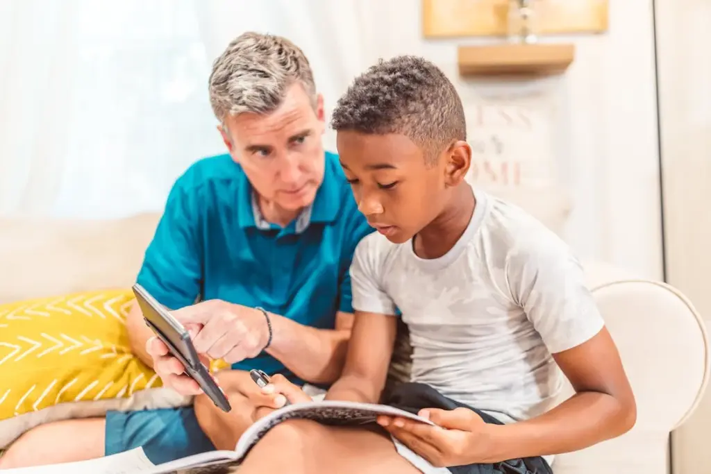 A man is assisting a boy with his homework on a couch. The man points to a tablet while the boy looks at a book.