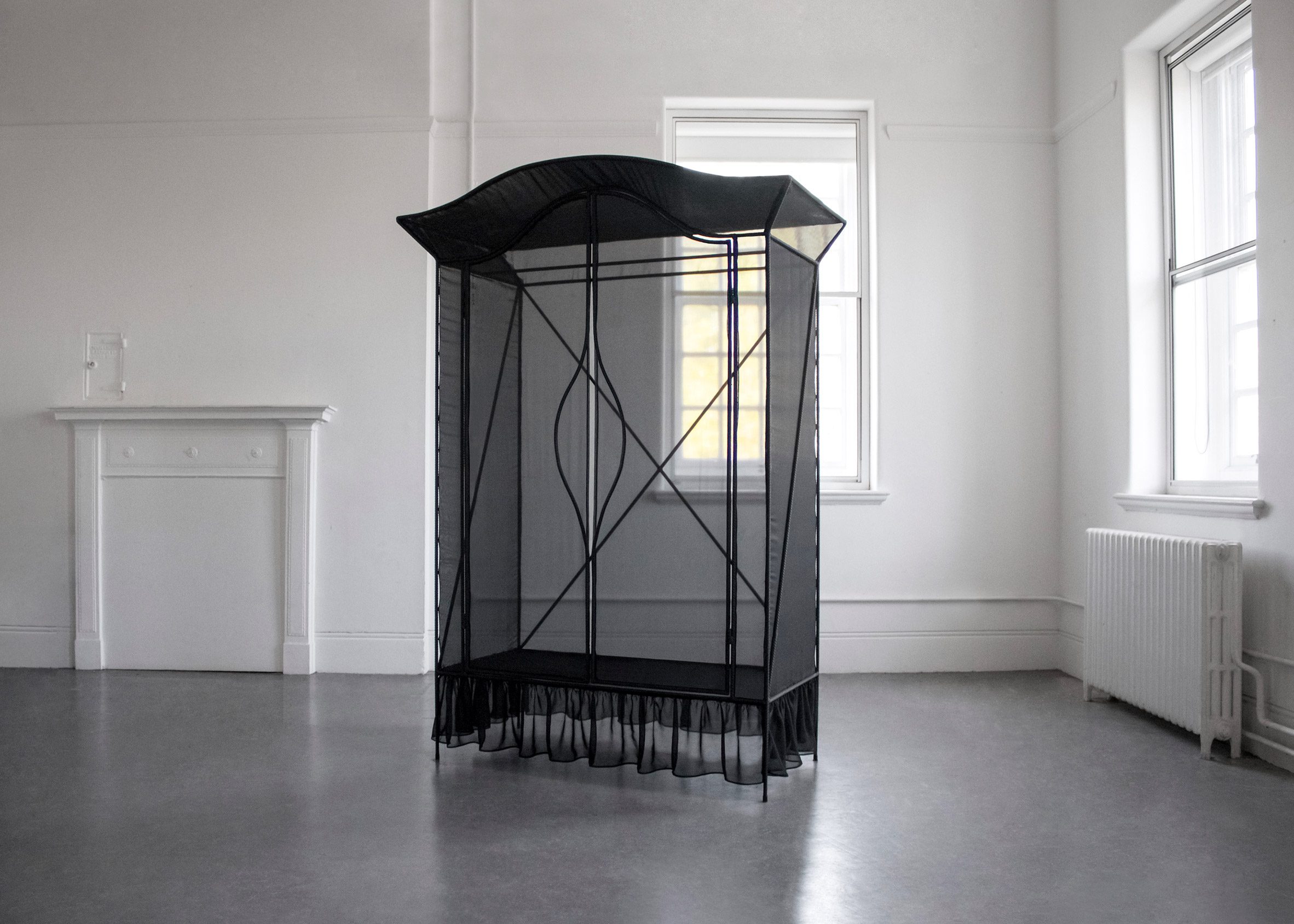 A photograph of a black wardrobe frame covered in black netting in a white room.