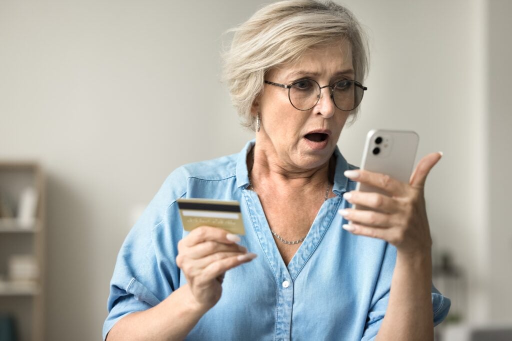 An older woman with glasses looks surprised at her smartphone while holding a credit card in her other hand.