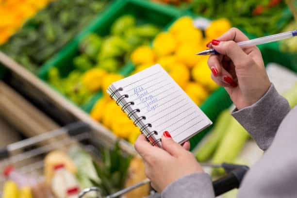 Person holding a pen and a grocery list in a notebook, standing in front of green crates filled with various vegetables and fruits in a supermarket.