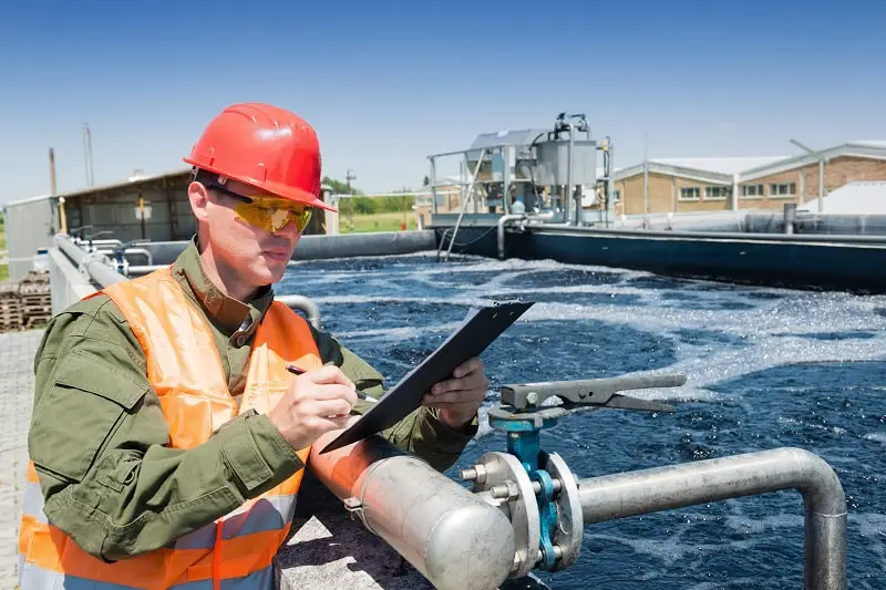 A worker in a safety vest and hard hat inspects a clipboard near an industrial water treatment facility, with large tanks and machinery in the background.