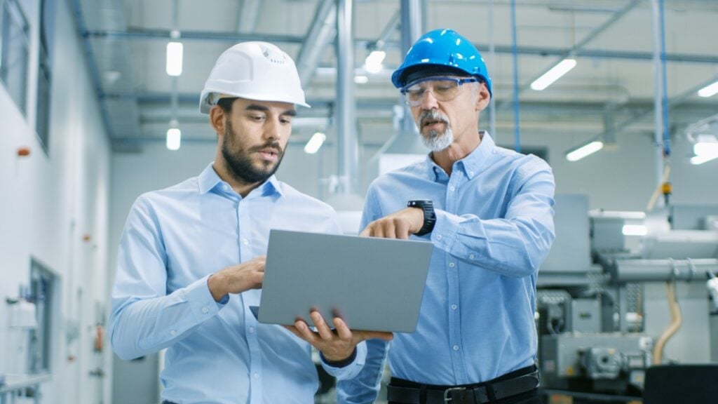 Two men in safety helmets stand in an industrial setting examining a laptop.