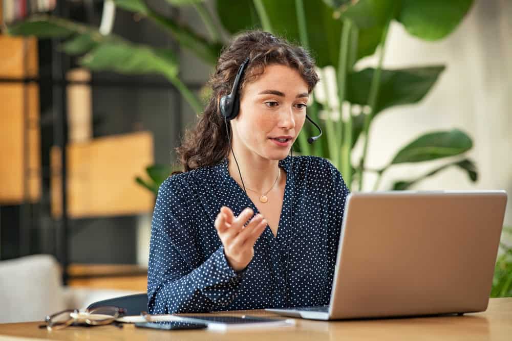 A stay-at-home mom wearing a headset sits at a desk, speaking while looking at a laptop screen. Green plants are in the background as she balances her role with stay-at-home jobs.