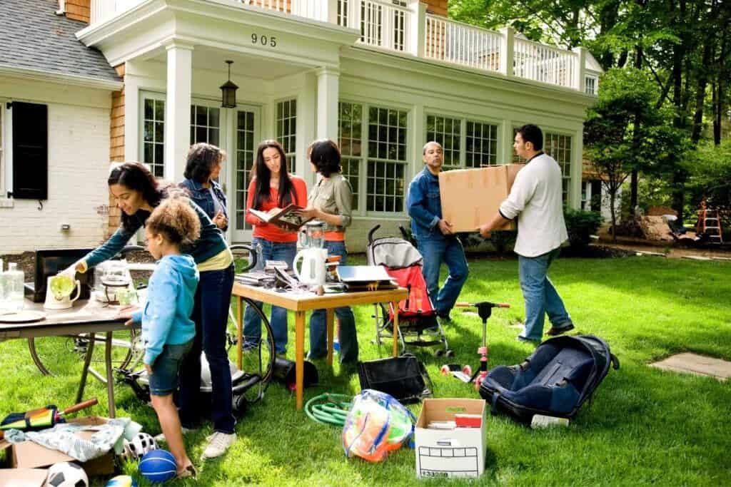 People organizing items on a table and carrying a box in front of a house with a lawn and garden.