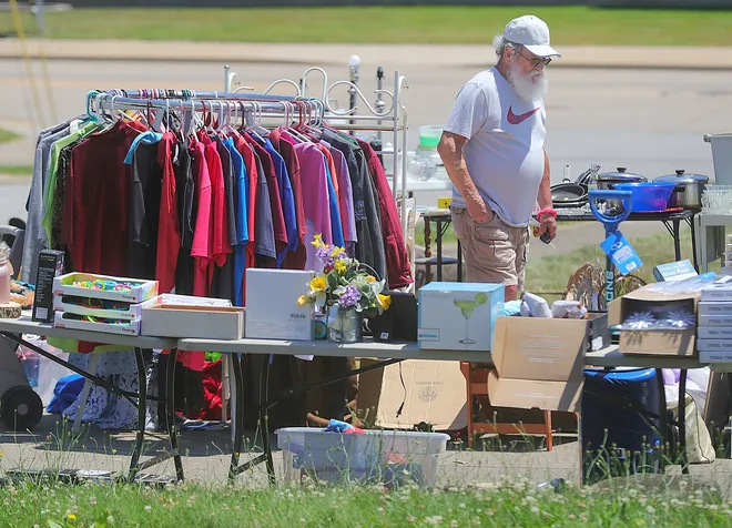 Man standing near a garage sale setup with clothing racks, various items, and flowers on tables.