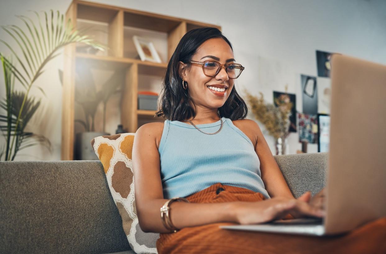 Smiling mixed race woman browsing internet on a laptop at home. One happy hispanic entrepreneur sitting alone on her sofa and blogging over a weekend. Relaxing in a living room and typing a blog post