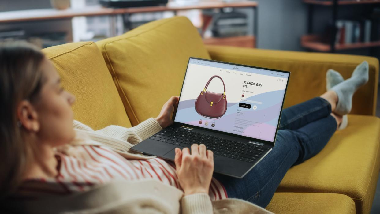 Beautiful Caucasian Female is Using Laptop Computer with Clothing Online Web Store to Choose and Buy Handbag from New Collection. Female Surfing the Net and Lying on Couch Sofa at Home Living Room.