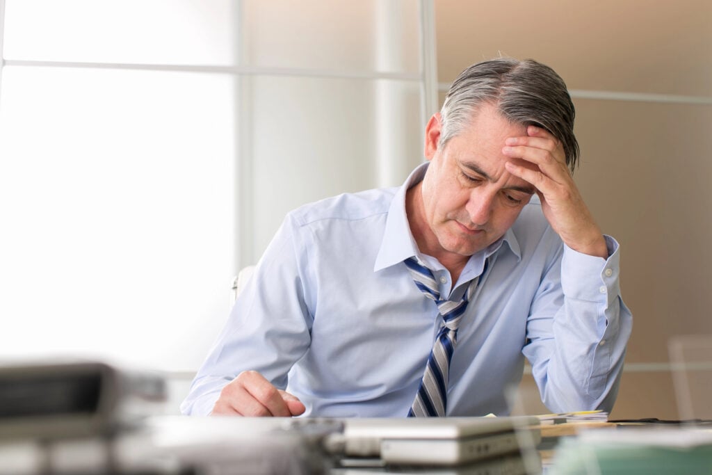 A man in a light blue shirt and striped tie sits at a desk, resting his head in his hand while looking down at paperwork, seemingly pondering why companies do not hire over 50.