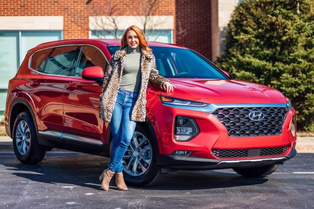 A woman standing next to a red hyundai santa fe suv.