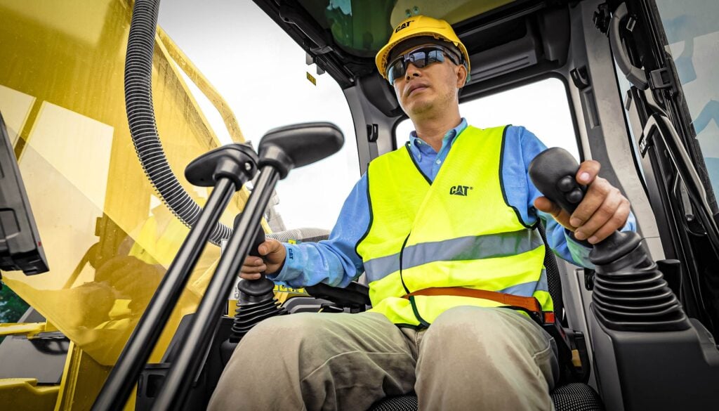 Person operating heavy machinery while wearing a hard hat, safety vest, and sunglasses inside the cabin.