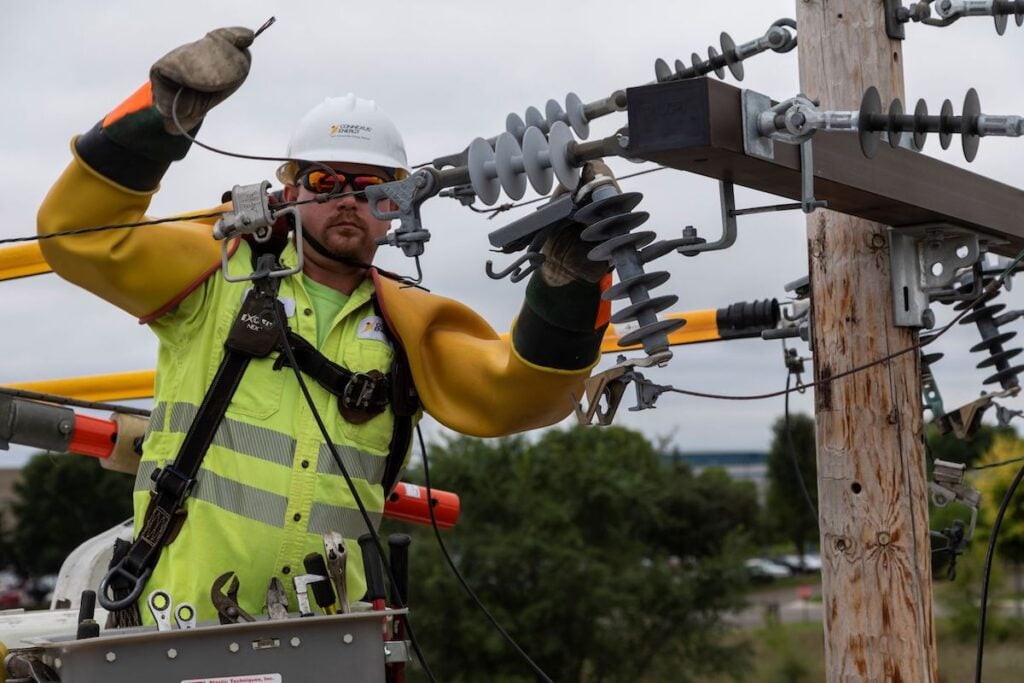 A utility worker in protective gear is repairing a power line on a pole.