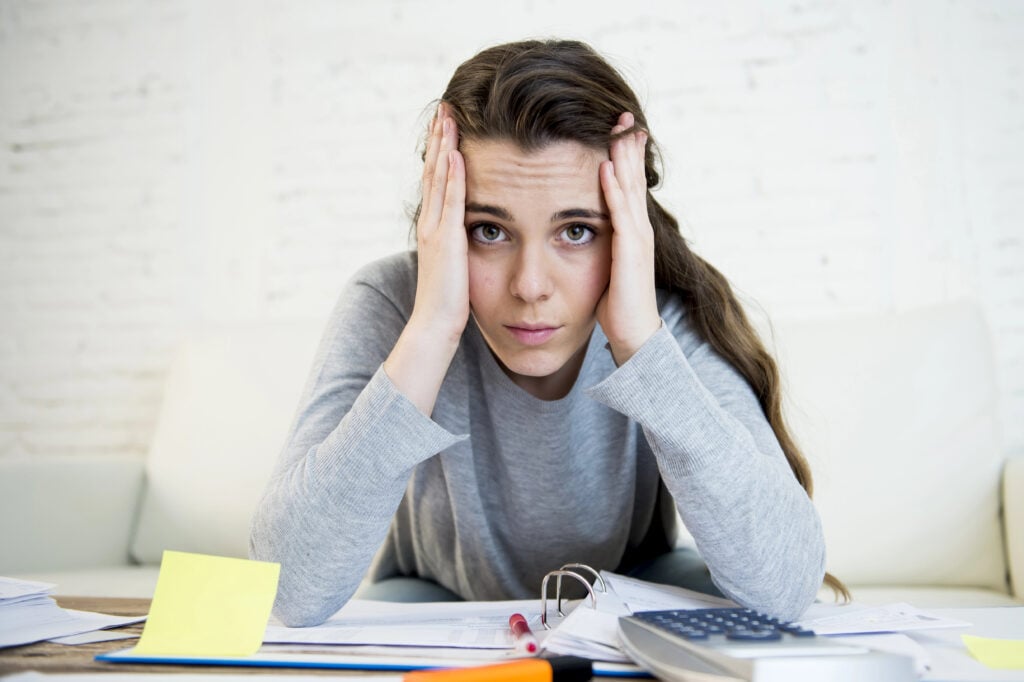 A woman sits at a table with papers, a binder, sticky notes, and a calculator, holding her head with both hands, looking stressed.