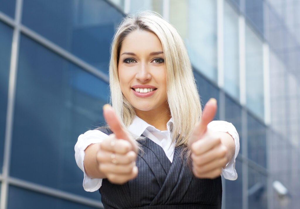 A smiling woman with blonde hair gives two thumbs up in front of a modern glass building. She is wearing a white shirt and a gray vest.