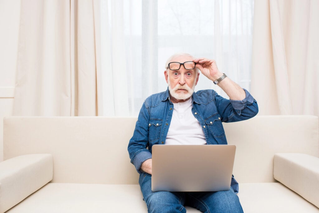 Elderly man with white beard and glasses, sitting on a beige sofa, looking surprised at a laptop screen.