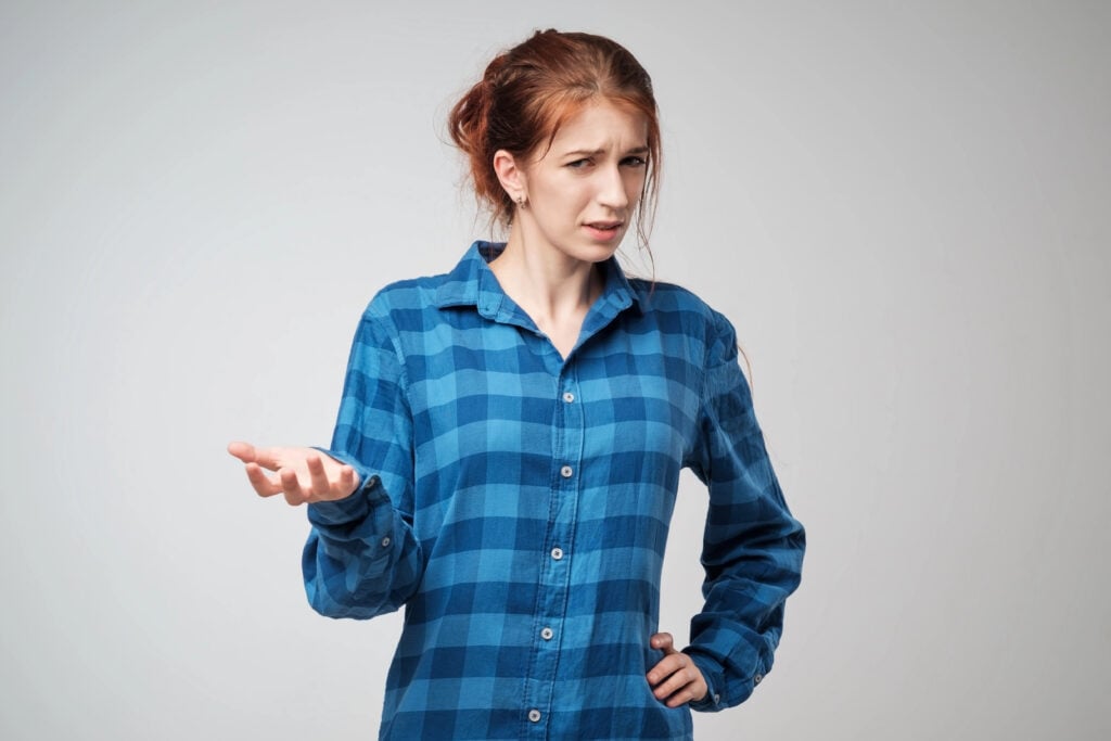 A person with red hair in a blue plaid shirt gestures with one hand, appearing confused or questioning, against a plain background.