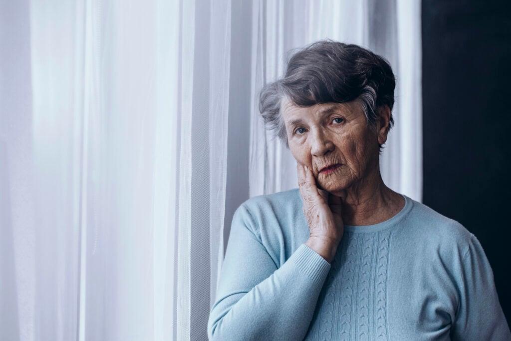 Elderly woman with short gray hair stands by a sheer curtain, resting her cheek in her hand and looking thoughtfully ahead. She wears a light blue sweater.