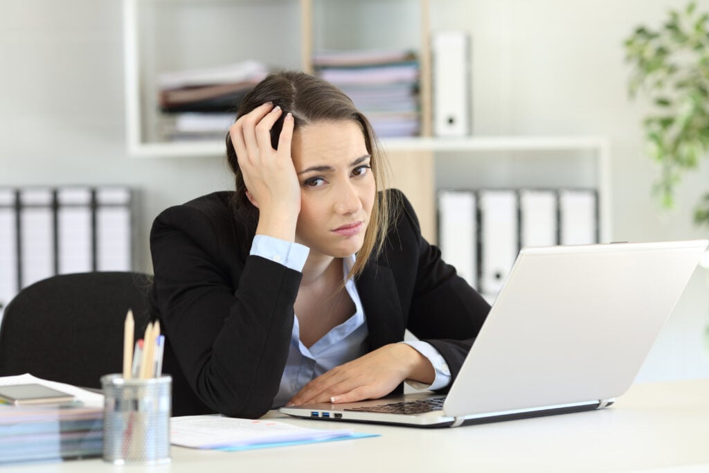 A woman in a business suit sits at a desk, looking stressed with her head resting on her hand, in front of an open laptop. Office supplies and file folders are visible in the background.