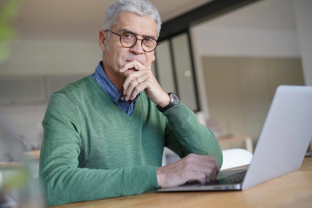 Older man with glasses and gray hair, wearing a green sweater, sits at a table with a laptop, resting his chin on his hand while looking thoughtfully at the camera.