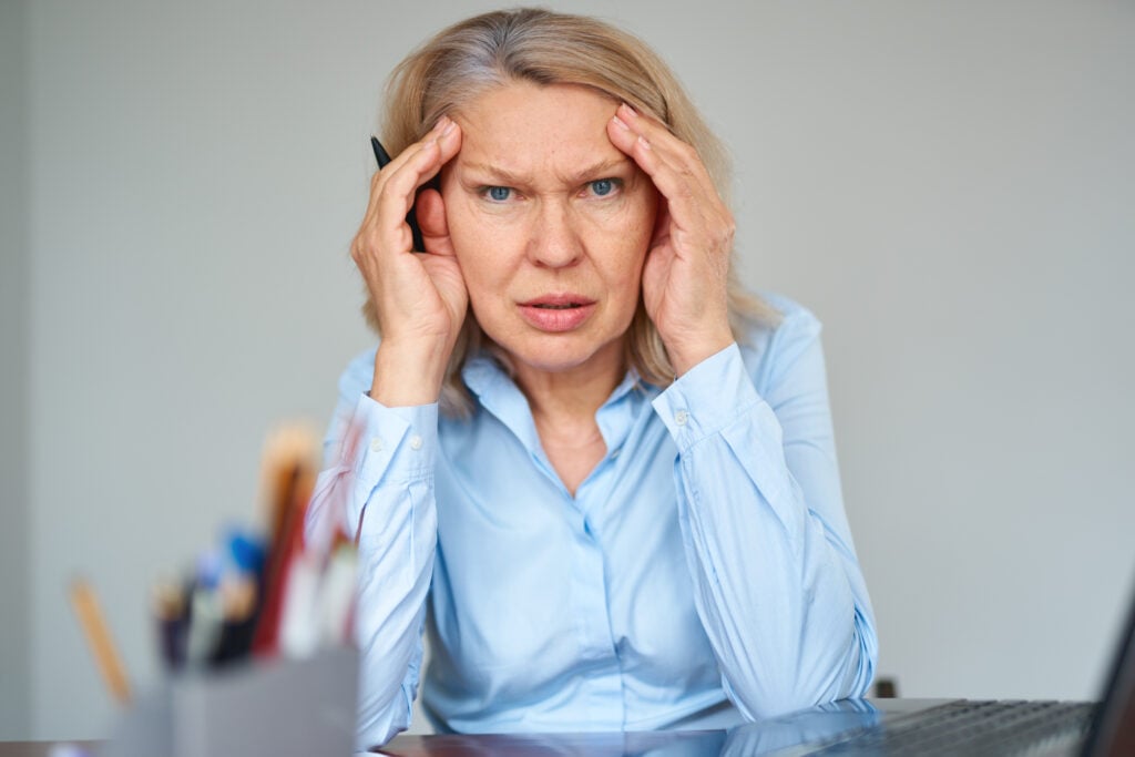 A woman with blonde hair wearing a blue shirt sits at a desk with her hands on her temples, looking concerned or stressed. A pencil cup with various items and part of a laptop are visible in the foreground, as she ponders why companies do not hire over 50.