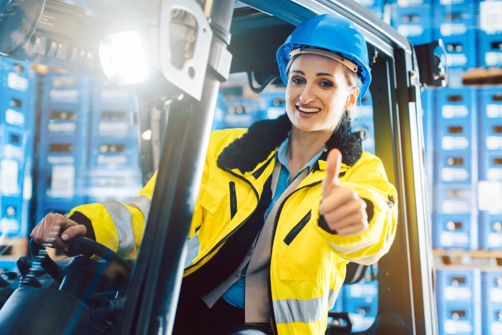 A person in a high-visibility jacket and blue hard hat operates a forklift inside a warehouse, giving a thumbs-up.